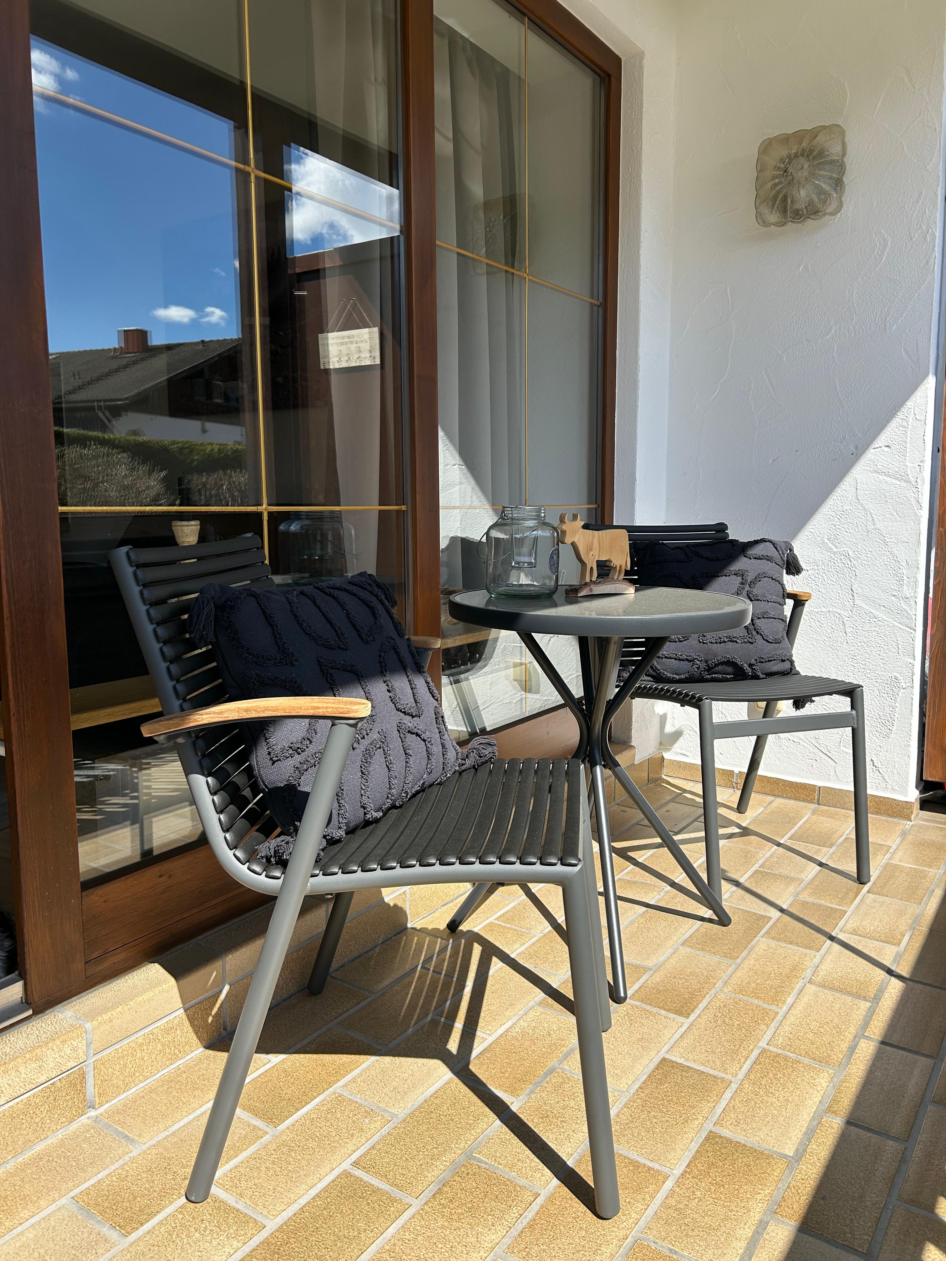 Sonniger Balkon der Ferienwohnung Sonnenstüble in Bad Hindelang mit zwei modernen Stühlen, schwarzen Kissen und einem kleinen runden Tisch vor der großen Glasfront