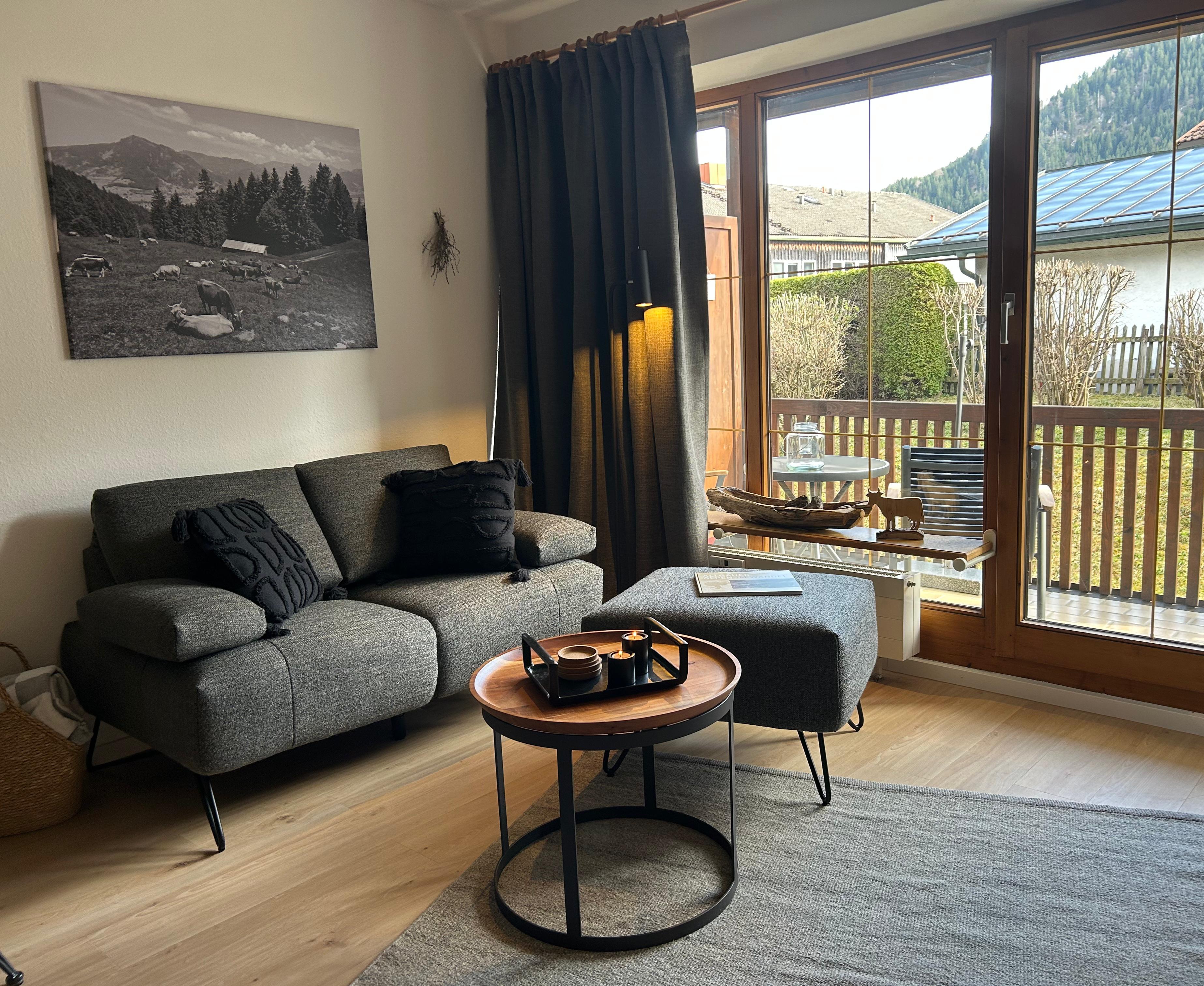 Gemütlicher Wohnbereich der Ferienwohnung Sonnenstüble mit grauem Sofa, Holztisch, Sessel und Blick durch das große Fenster auf die Terrasse in Bad Hindelang