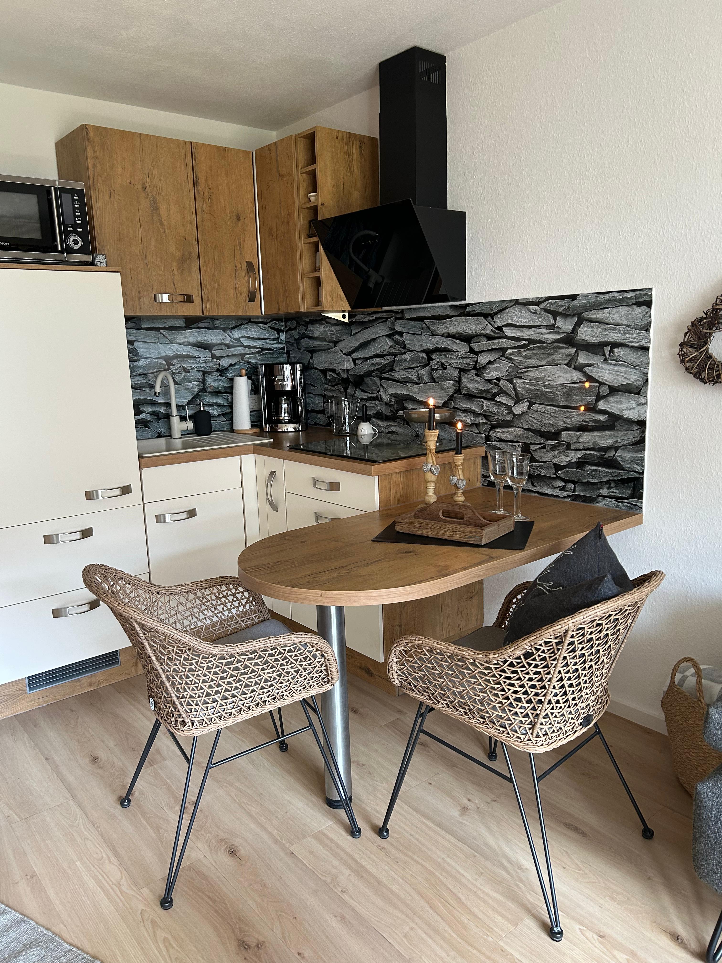 Essecke der Ferienwohnung mit rundem Holztisch, zwei Korbstühlen und angrenzender, modern ausgestatteter Küche vor rustikaler Steinwand
