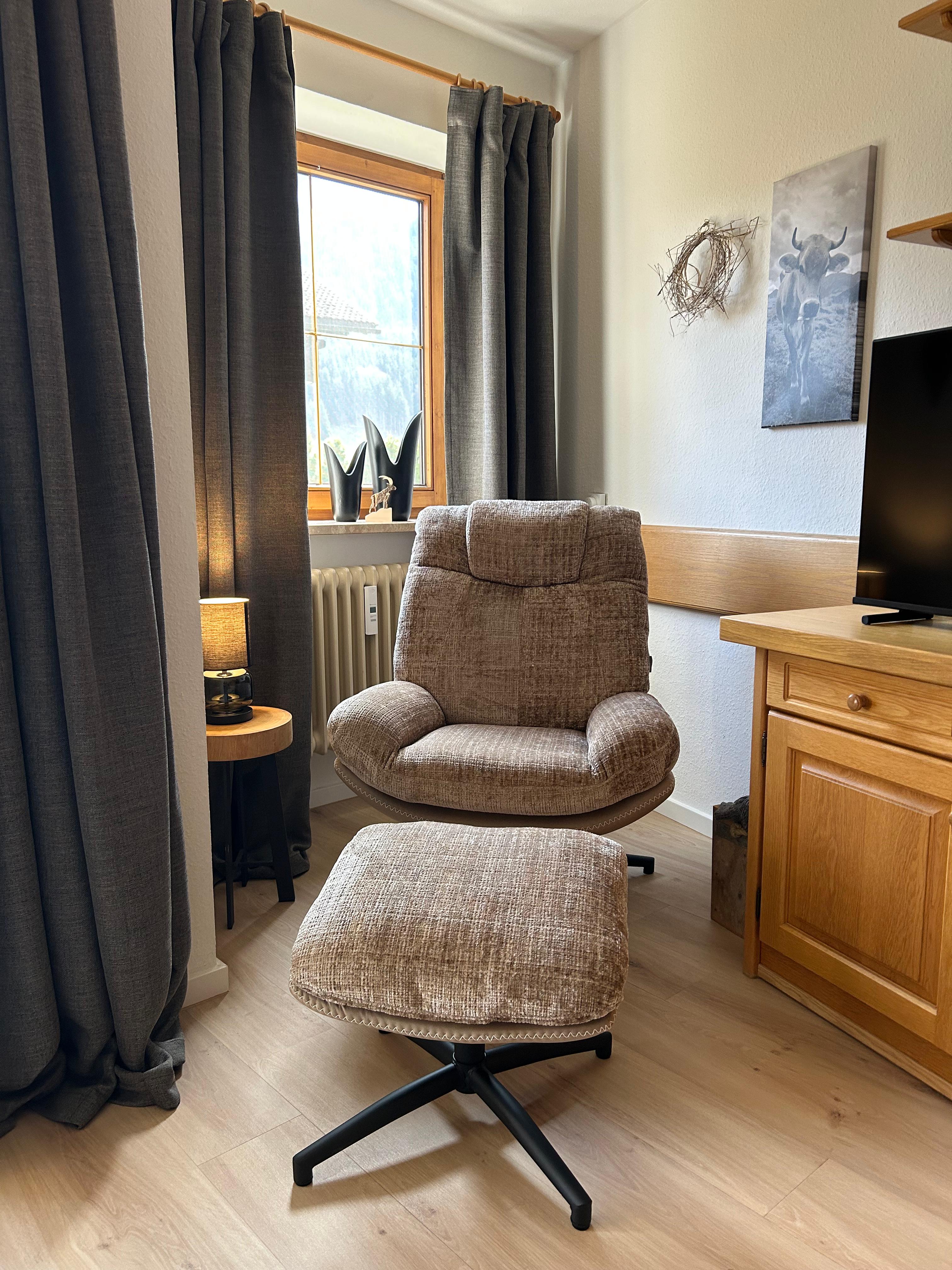 Heller Wohnraum im Allgäuer Stil mit bequemem Fernsehsessel, Fußhocker, Massivholz-Sideboard mit TV und Fensterausblick